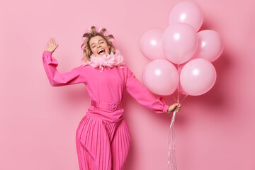 Canvas Print - People fun and celebration concept. Carefree glad woman wears long dress applies hair rollers celebrates birthday holds bunch of helium balloons isolated over pink background dances with joy