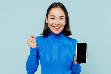 Wall Mural - Fun young woman of Asian ethnicity 20s wears blue shirt hold use mobile cell phone with blank screen workspace area doing winner gesture isolated on plain pastel light blue background studio portrait.