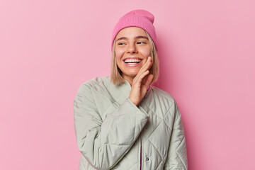 Wall Mural - Joyful carefree teenage girl with happy expression smiles positively keeps hand on cheek being in good mood wears hat and jacket isolated over pink studio background. Positive emotions concept