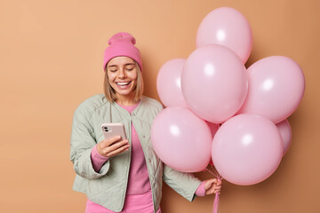 Canvas Print - Happy smiling young European woman has good festive mood hods bunch of inflated balloons uses mobile phone chats with friends wears hat and jacket comes on party isolated over brown background