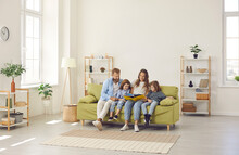 Happy Family Spending Time At Home. Mum, Dad And Little Kids Reading Book While Sitting Together On Green Sofa In Room With Light Walls, Beige Rug And Simple Brown Shelves In Their Big New House