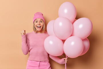 Wall Mural - Joyful woman clenches fist and celebrates good news celebrates special occasion poses wth inflated balloons dressed in pink clothes poses against brown background. People and festivity concept