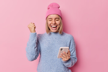 Excited overjoyed woman holds mobile phone and does winner gesture celebrates winning lottery triumphs over success dressed in casual jumper and hat isolated over pink background. Yes I did it