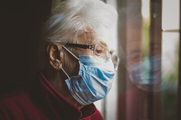 Wall Mural - Old woman portrait at the window. Grandmother with medical mask staying home alone during coronavirus pandemic quarantine