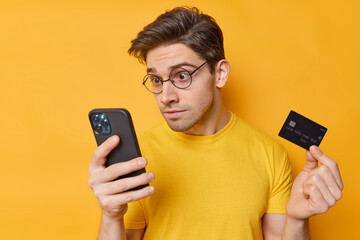 Impressed stunned brunet man stares at smartphone screen holds credit card reacts on unexpected discounts wears round spectacles casual t shirt isolated over yellow background downloads app.