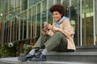 © wayhome.studio - Outdoor shot of happy curly haired millennial girl wears fashionable clothes sits on stairs near modern shopping centre reads message on smartphone enjoys recreation time. Modern technologies