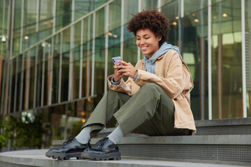 Wall Mural - outdoor shot of happy curly haired millennial girl wears fashionable clothes sits on stairs near mod