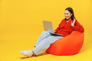 Wall Mural - Full size young woman of Asian ethnicity 20s in casual clothes sit in bag chair hold work on laptop pc computer get video call talk greet with hand isolated on plain yellow background studio portrait.