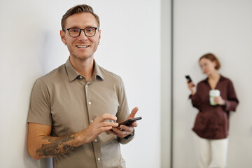 Wall Mural - Minimal waist up portrait of smiling tattooed man holding smartphone and looking at camera in office, copy space