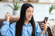 © kucherav - Excited Asian young woman holding a smartphone in her hands, looking at the screen of a mobile phone and smilling. Excited of getting win news, rejoices at good news, a big win in the lottery