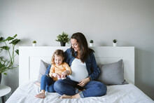 Happy Pregnant Woman Looking At Daughter Holding Baby Booties On Bed