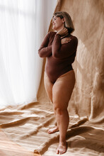 Smiling Curvy Woman Wearing Brown Bodysuit Standing In Front Of Backdrop