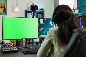 Focused player woman playing online videogames during gaming tournament loooking at mok up green screen chroma key computer with isolated display. Gamer streaming space shooter game using joystick