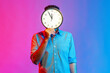 Portrait of man in shirt hiding face behind big wall clock display, wasting his time, procrastination, bad organization of working time. Indoor studio shot isolated on colorful neon light background.