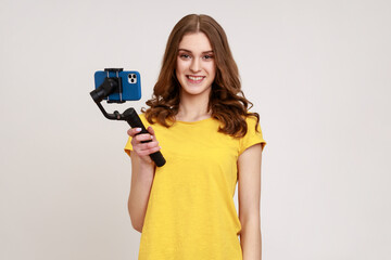 Poster - Portrait of blogger teenager girl in yellow T-shirt shoots video for vlog on electronic stabilizer, holding steadicam. looking smiling at camera. Indoor studio shot isolated on gray background.