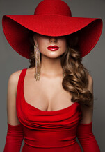 Fashion Woman In Hat Hidden Face In Red Decollete Dress With Golden Earring. Elegant Lady In Plunging Neckline Evening Gown. Sexy Model Portrait With Glossy Red Lips Over Gray Studio Background