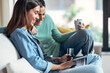 © nenetus - Two smiling young women talking while paying something online with her credit card sitting on couch at home.
