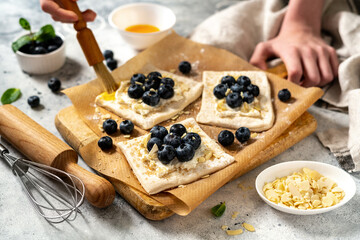 Wall Mural - Raw puff pastry with cheese cream and blueberries. The process of making delicious sweet buns or mini pies. Greasing the edges of the baking dough with egg. Hands in the frame