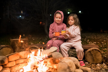 Fun camping activities for kids. Girls sitting by crackling campfire and drinking hot chocolate in the evening. Family getaway and hiking in woodland camp with children