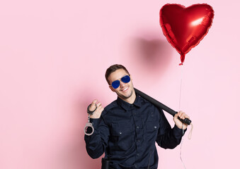 Young charming policeman officer with heart shape balloon wearing sunglasses isolated on pink background. St, Valentine's day