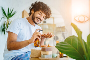 Happy hansome latin man holding glass bottle with dispenser and take care of his skin in home clothes relaxing on bathroom and smiling.