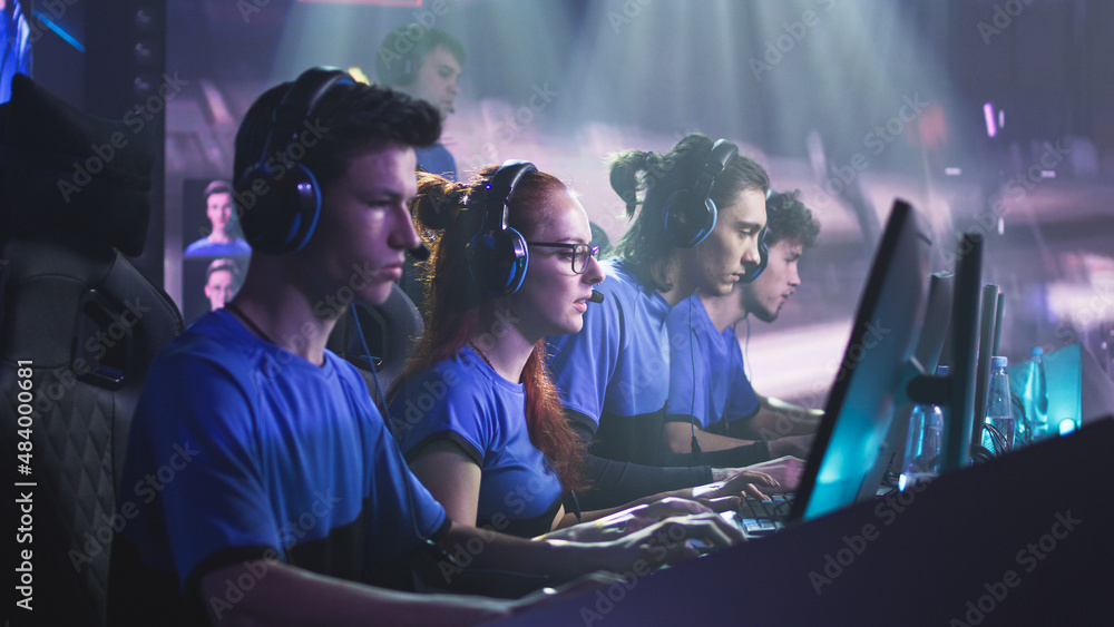 Young and successful men and women professional gamers with coach playing video game on computers against monitor with match broadcast during esports tournament