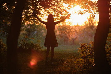 Dark silhouette of young woman walking alone through dark forest joyfully raising her hands in summer evening. Enjoying nature and outdoor activities concept