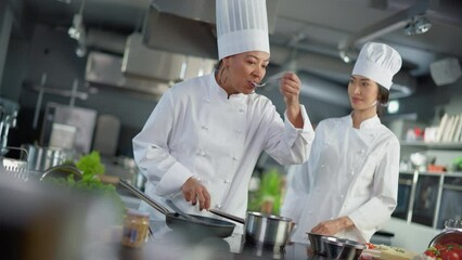 Wall Mural - Restaurant Kitchen: Portrait of Asian and Black Female Chefs Preparing Dish, Tasting Food, Doing High-Five in Successful Celebration. Two Professionals Cooking Delicious, Authentic Food, Healthy Meals