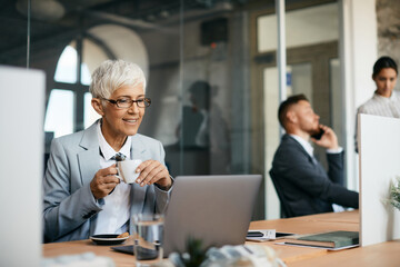 Senior businesswoman enjoys in cup of coffee while using laptop in the office.