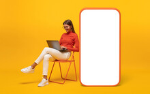 Happy Young Woman Sitting With Laptop On Chair Near Blank Screen Of Huge Phone, Mockup For App