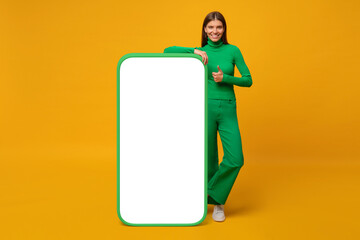 Happy young woman standing and showing blank screen of huge phone, mock up for your app