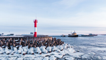 Wall Mural - Red-white lighthouse standing on island surrounded by frozen breakwaters against cargo ships sailing on clear water surface under blue sky in winter