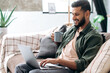© Kateryna - Positive handsome young indian man in casual clothes, sitting on the couch at home in the living room, using a laptop, browsing the Internet, messaging on social networks, looking at screen, smiles