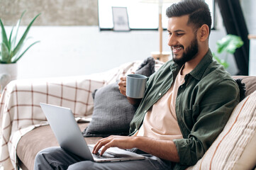 Positive handsome young indian man in casual clothes, sitting on the couch at home in the living room, using a laptop, browsing the Internet, messaging on social networks, looking at screen, smiles