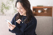 © lielos - ソファーに座りスマートフォンを見ている日本人女性、自宅リビング