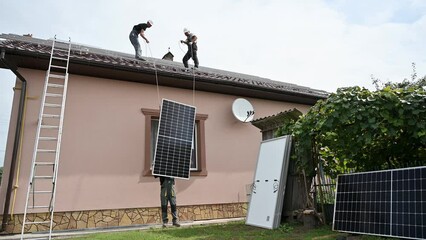 Wall Mural - Men workers lifting up photovoltaic solar modul on roof of house. Electricians in helmets installing solar panel system outdoors. Concept of alternative and renewable energy.