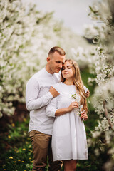 Wall Mural - A happy young couple in love stands in a garden of blooming apple trees. A man in a white shirt and a girl in a white light dress are walking in a flowering park