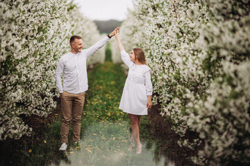 Wall Mural - Happy family couple in spring blooming apple orchard. Young couple in love enjoy each other while walking in the garden. The man holds the woman's hand. Family relationships