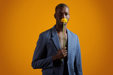 Wall Mural - Young African man with yellow flower by his face standing in front of camera against orange background and looking at you