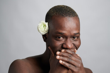 Wall Mural - Young African man with white camelia flower by ear covering his mouth while laughing in front of camera in isolation