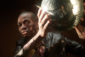 Wall Mural - Serious young African man in leather jacket holding discoball and looking at you while peeking out of it in the night club