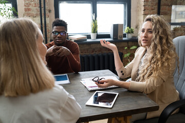 Poster - Young confident businesswoman in formalwear consulting female colleague while sitting by table between intercultural co-workers