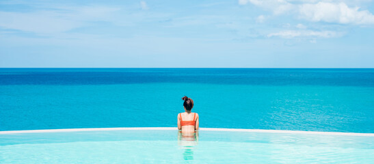 Happy woman in orange swimsuit swimming in infinity pool at luxury hotel against ocean front. young female enjoy in tropical resort. Relaxing, summer,  travel, holiday, vacation and weekend concept