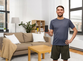 Wall Mural - fitness, sport and healthy lifestyle concept - smiling man in sports clothes showing smartphone over home room background