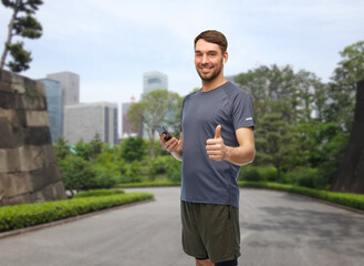 Sticker - fitness, sport and healthy lifestyle concept - smiling man in sports clothes with smartphone and earphones listening to music and showing thumbs up gesture over park in tokyo city, japan on background