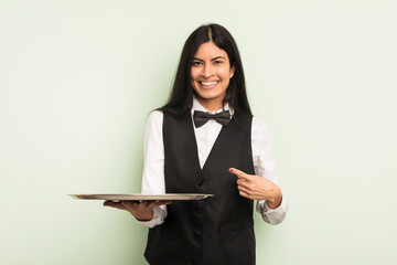 Wall Mural - young pretty hispanic woman smiling cheerfully, feeling happy and pointing to the side. waiter with tray concept