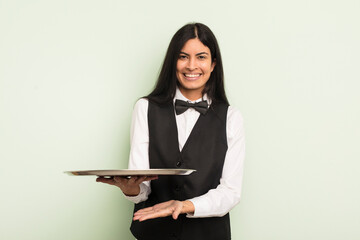 Wall Mural - young pretty hispanic woman smiling cheerfully, feeling happy and showing a concept. waiter with tray concept