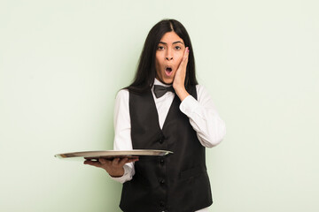 Wall Mural - young pretty hispanic woman feeling shocked and scared. waiter with tray concept