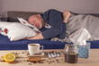 Mężczyzna przeziębiony leży z gorączką w łóżku na pierwszym planie widzimy tabletki, herbatę, miód, cytrynę i korzenie pomocne przy leczeniu.