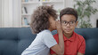 Little black girl whispering secret to brother in eyeglasses, siblings gossiping